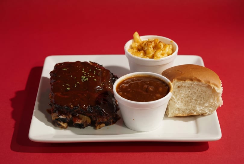 BBQ ribs with sauce, baked beans, mac and cheese, and a dinner roll on a white plate.