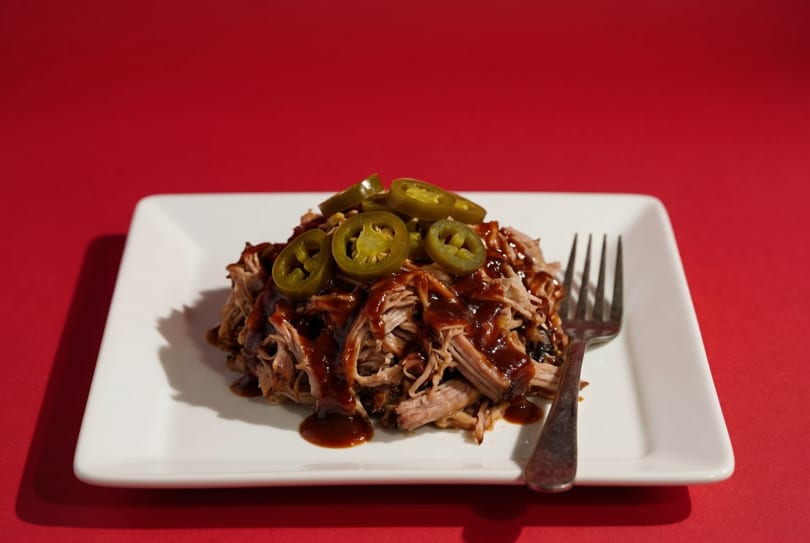Pulled pork with barbecue sauce and sliced jalapeños on a white rectangular plate with a fork.