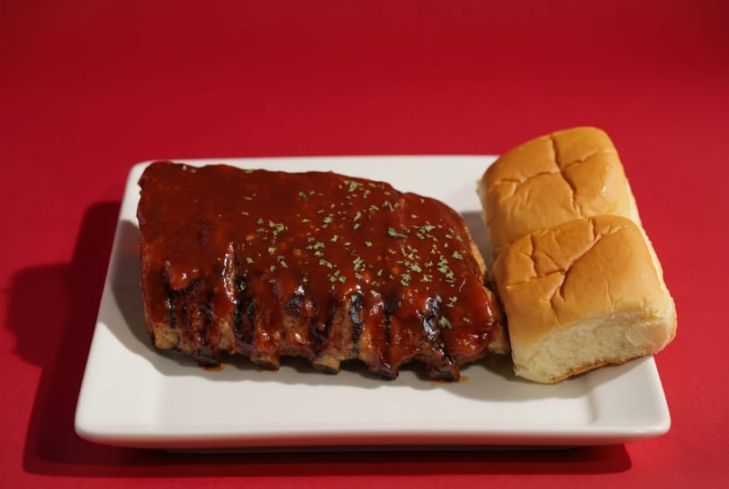 A rack of BBQ ribs with sauce and parsley, served with two dinner rolls on a white plate.