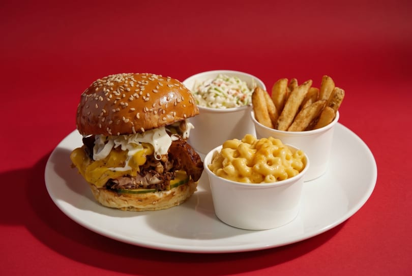 A burger with pulled pork, cheese, and coleslaw, served with fries, mac and cheese, and coleslaw.