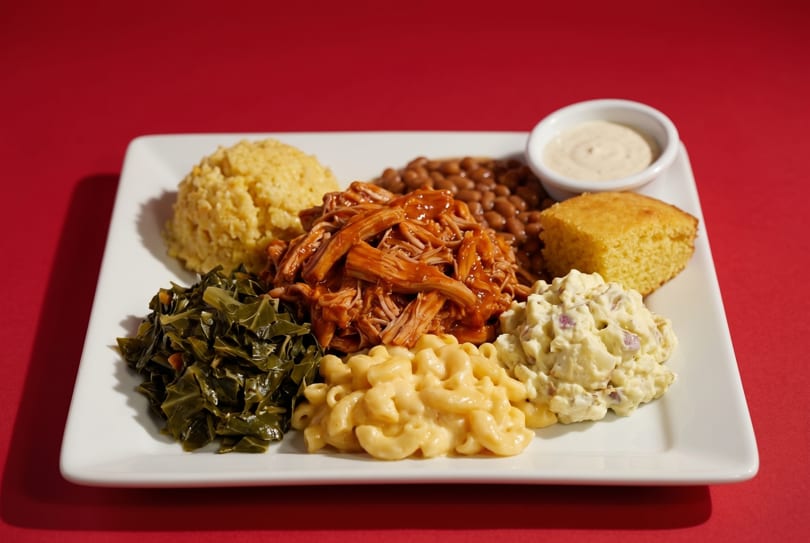 A plate of BBQ pulled pork, collard greens, mac and cheese, potato salad, baked beans, cornbread, and grits.