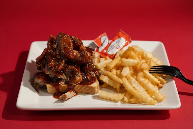 BBQ chicken sandwich with fries and ketchup packets on a white plate against a red background.